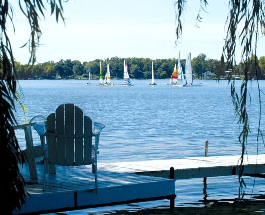 An adirondack chair sits on a dock looking out on sailboats on a lake.